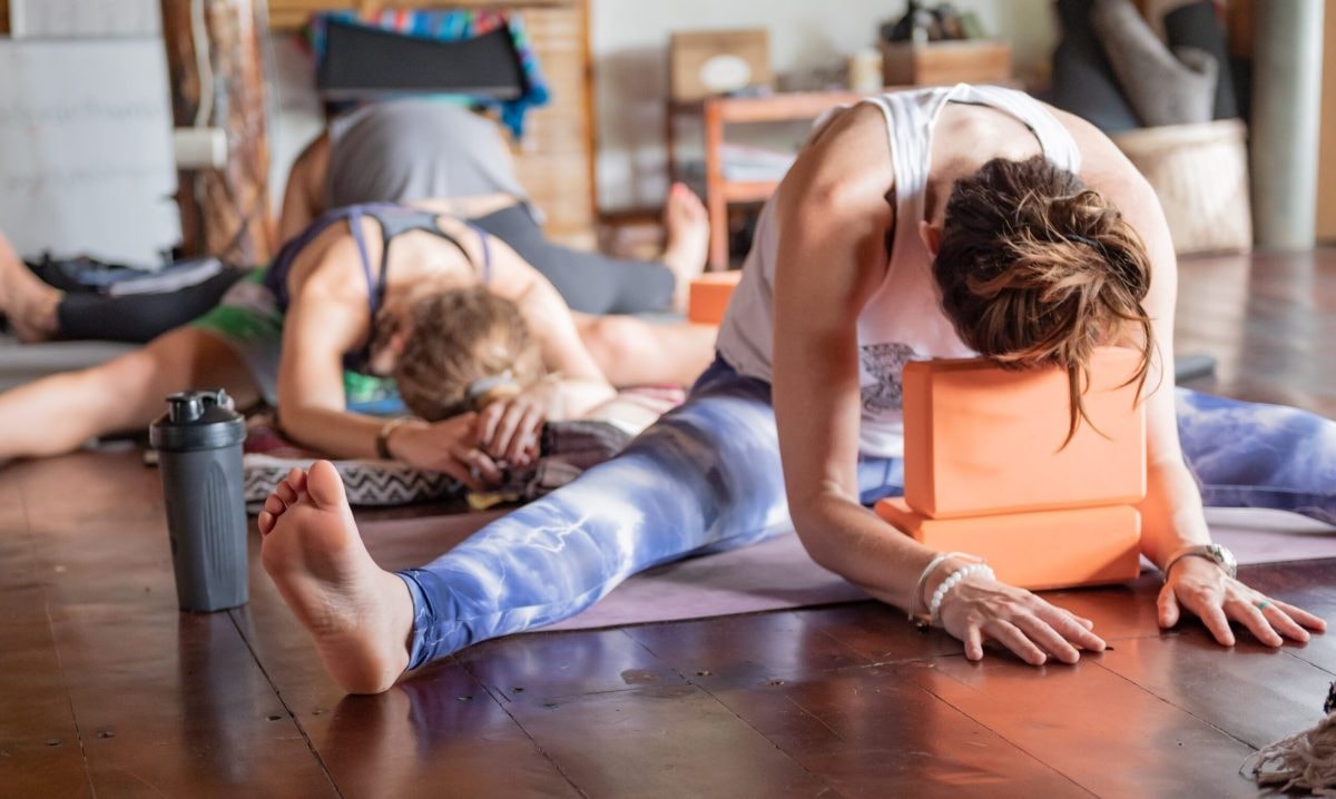 Two students in a seated forward fold on the wooden floor of the shala