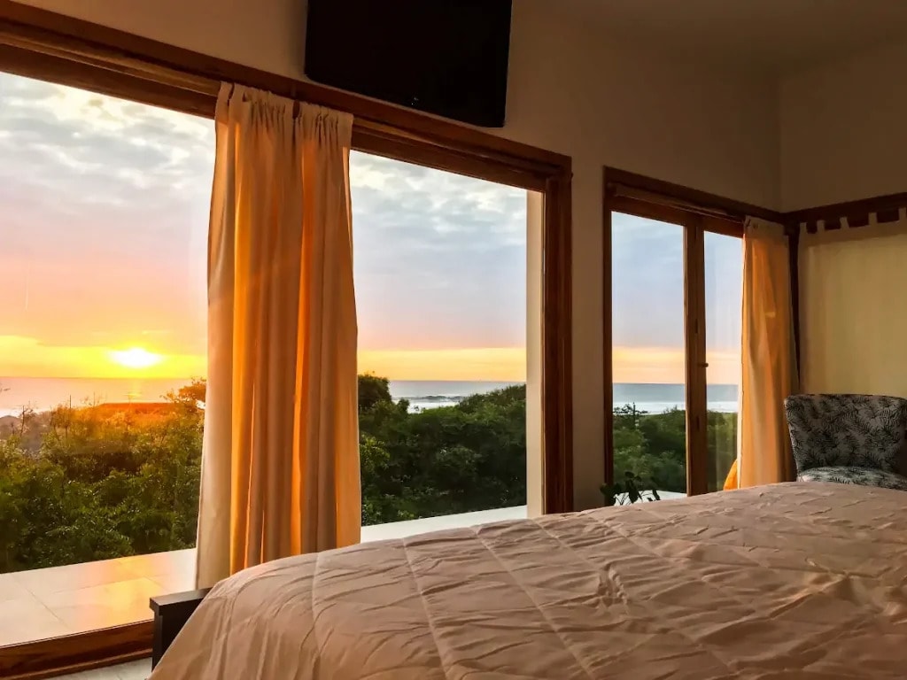 A bed facing open curtains and the ocean at sunset