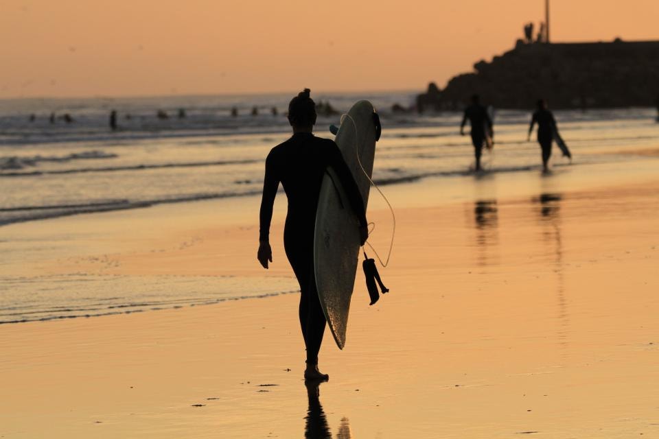 A surfer walking the sand at Imsouane at sunset with the lineup behind